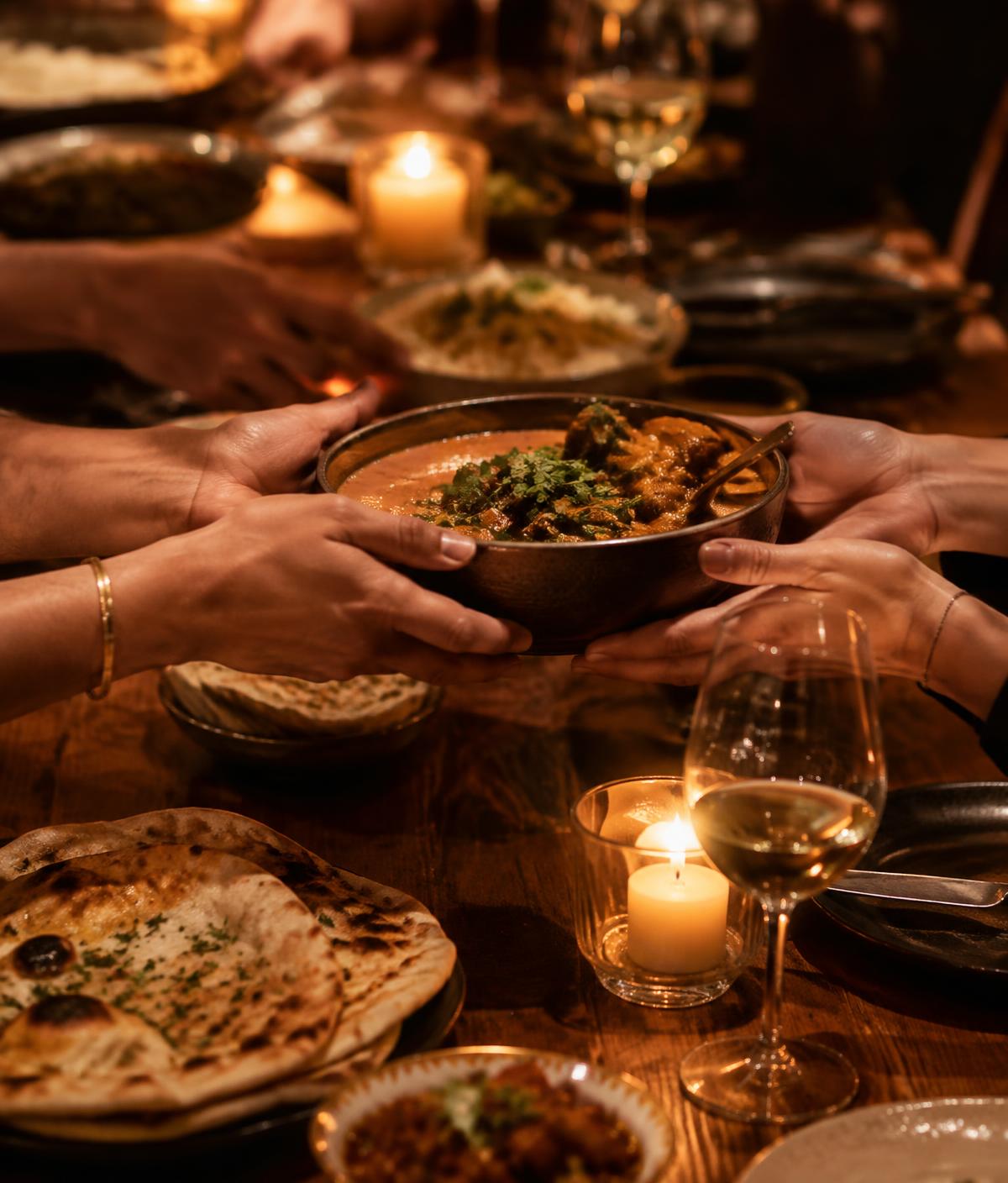 Hands passing a bowl of curry across a candlelit table set with naan and side dishes